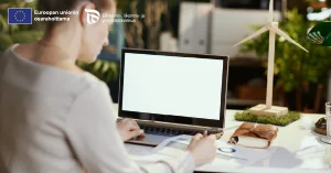 Henkilö työskentelee pöydän ääressä avoimen kannettavan tietokoneen kanssa, jonka näyttö on valkoinen; pöydällä on muistivihko, paperia ja pienen tuuliturbiinin malli, ja taustalla näkyy kasveja sekä EU:n osarahoitustunnus.