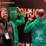 Kuvassa kaksi vihreisiin huppareihin pukeutunutta henkilöä seisoo tapahtumataustan edessä. Vasemmalla oleva henkilö pitää olkapään päällä vihreää ämpäriä, jossa lukee “VICTIS” ja pienempänä teksti vihreästä ICT:stä, sekä näyttää toisella kädellä rauhanmerkkiä. Oikealla oleva henkilö on selkä kameraan päin ja osoittaa peukaloillaan hupparin selkäpuolella olevaa kuvioita ja tekstiä “Green ICT -ekosysteemi”. Taustalla näkyy “SOHJO”-teksti. Yläreunassa ovat Euroopan unionin osarahoitusmerkintä sekä Elinvoimakeskuksen logo.