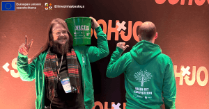 Kuvassa kaksi vihreisiin huppareihin pukeutunutta henkilöä seisoo tapahtumataustan edessä. Vasemmalla oleva henkilö pitää olkapään päällä vihreää ämpäriä, jossa lukee “VICTIS” ja pienempänä teksti vihreästä ICT:stä, sekä näyttää toisella kädellä rauhanmerkkiä. Oikealla oleva henkilö on selkä kameraan päin ja osoittaa peukaloillaan hupparin selkäpuolella olevaa kuvioita ja tekstiä “Green ICT -ekosysteemi”. Taustalla näkyy “SOHJO”-teksti. Yläreunassa ovat Euroopan unionin osarahoitusmerkintä sekä Elinvoimakeskuksen logo.